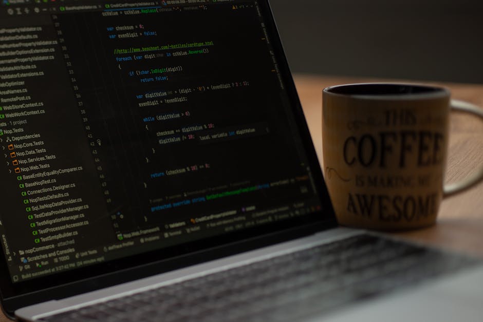 A close-up view of a laptop on a wooden desk displaying lines of computer code within a dark-themed programming interface. To the right of the laptop, there is a ceramic mug with a coffee-themed quote