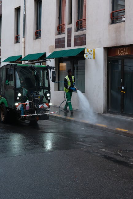 A female cleaner from Battersea Cleaners is performing surface cleaning in a living room, wiping down a wooden dining table with a cloth. She is wearing a black uniform with gloves and appears to be t